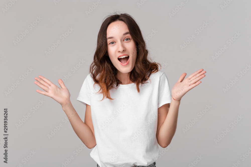 Excited brunette woman in a white tee shrugging her shoulders expressing emotion of surprise standing isolated over grey background. Emotion of surprise.