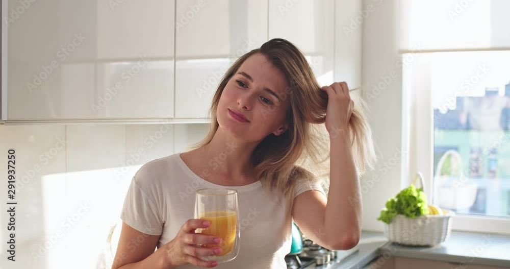 Portrait shot of the young beautiful Caucasian blonde woman looking straight to the camera and smiling joyfully with a glass of orange juice in the cozy light kitchen in the morning. Close up. Portrait shot of the young beautiful Caucasian blonde woman looking straight to the camera and smiling joyfully with a glass of orange juice in the cozy light kitchen in the morning. Close up.