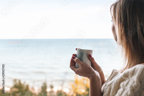 Side view portrait of a woman drinking coffee and looking outdoors through a window of an hotel room or home with the sea in the background