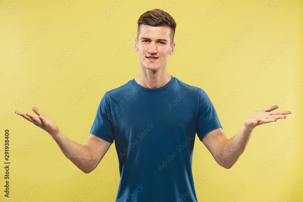 Caucasian young man's half-length portrait on yellow studio background. Beautiful male model in blue shirt. Concept of human emotions, facial expression. Showing and pointing something. Inviting.