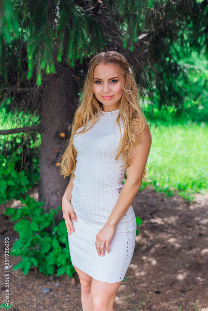 Portrait of a charming blond woman wearing beautiful white dress standing under fur tree. Portrait of a charming blond woman wearing beautiful white dress standing under fur tree.