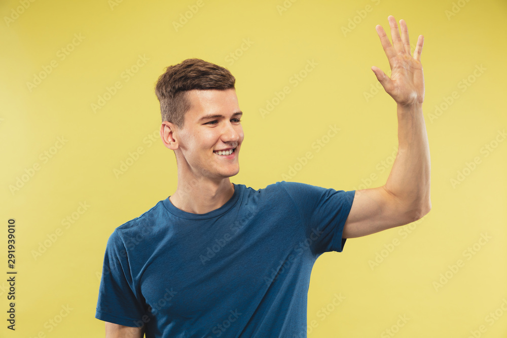 Caucasian young man's half-length portrait on yellow studio background. Beautiful male model in blue shirt. Concept of human emotions, facial expression. Greeting to somebody, smiling, confident. Caucasian young man's half-length portrait on yellow studio background. Beautiful male model in blue shirt. Concept of human emotions, facial expression. Greeting to somebody, smiling, confident.