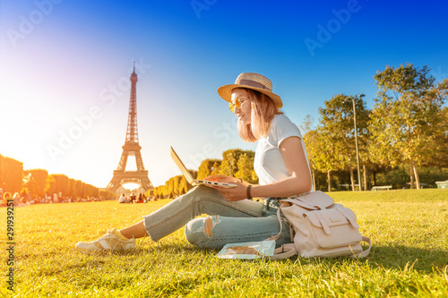 An Asian girl works on a laptop sitting on the grass on the Champ de Mars in Paris overlooking the Eiffel tower at sunset. Concept of remote work and freelancing © EdNurg