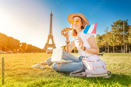 Happy Asian girl with French flag eating croissant and learning foreign languages on laptop with Eiffel tower in the background. The concept of modern distance learning for students in Paris © EdNurg