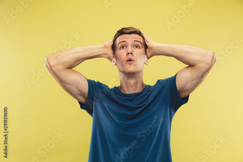 Caucasian young man's half-length portrait on yellow studio background. Beautiful male model in blue shirt. Concept of human emotions, facial expression. Looking up, looks shocked, astonished. © master1305