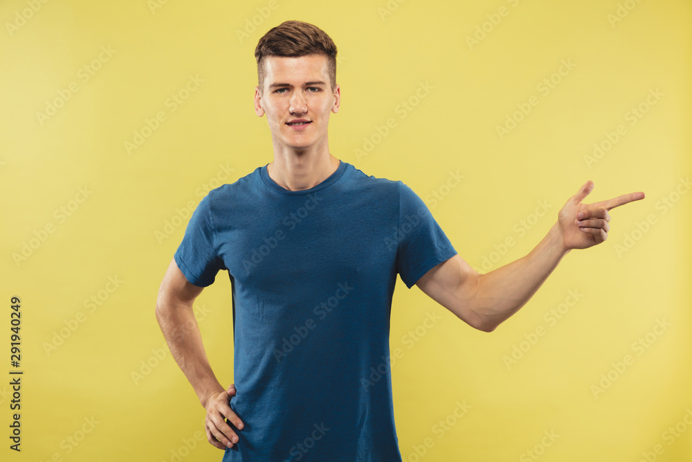 Caucasian young man's half-length portrait on yellow studio background. Beautiful male model in blue shirt. Concept of human emotions, facial expression. Showing and pointing something. Inviting. Caucasian young man's half-length portrait on yellow studio background. Beautiful male model in blue shirt. Concept of human emotions, facial expression. Showing and pointing something. Inviting.