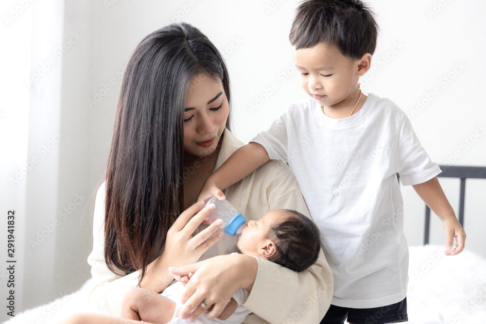 Young Asian mother hugging a cute newborn baby and feeding milk form bottle in with the son white bedroom. Duty of the wife to raise children. Mother and child concept Young Asian mother hugging a cute newborn baby and feeding milk form bottle in with the son white bedroom. Duty of the wife to raise children. Mother and child concept