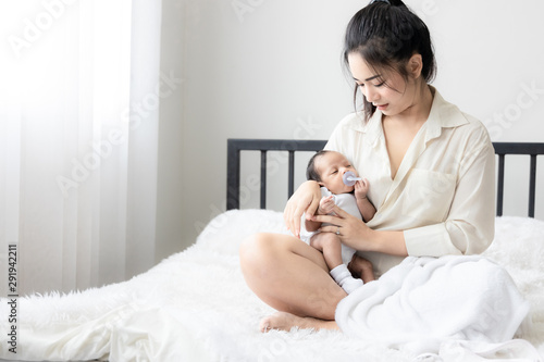 Portrait of Asian young mother with cute newborn baby sucking fake nipples and looking hers mother in white bedroom. Duty of the wife to raise children. Mother and child concept