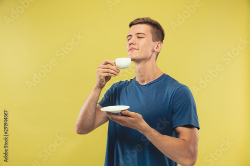 Caucasian young man's half-length portrait on yellow studio background. Beautiful male model in blue shirt. Concept of human emotions, facial expression. Enjoying drinking coffee or tea.
