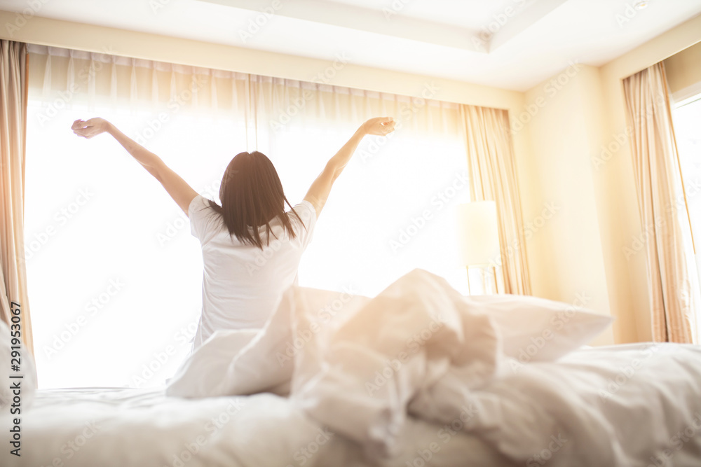 Woman stretching in bed after waking up, back view. Woman sitting near the big white window while stretching on bed after waking up with sunrise at morning, back view. Woman stretching in bed after waking up, back view. Woman sitting near the big white window while stretching on bed after waking up with sunrise at morning, back view.