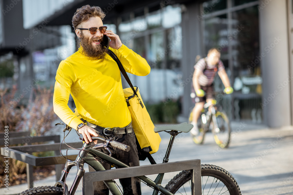 Young bearded courier delivering food in thermal bag on a bicycle, talking phone with a client on the city street Young bearded courier delivering food in thermal bag on a bicycle, talking phone with a client on the city street