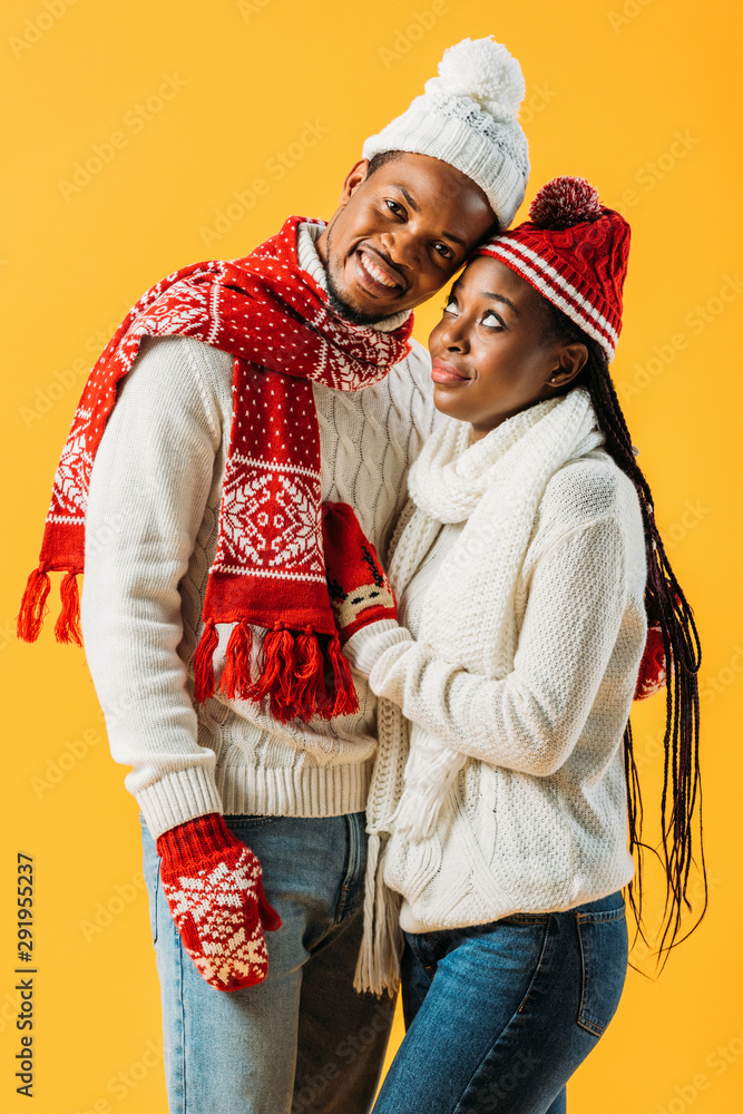 African American man in winter outfit embracing woman who looking at him isolated on yellow
