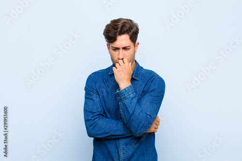 young handsome man feeling serious, thoughtful and concerned, staring sideways with hand pressed against chin against blue background