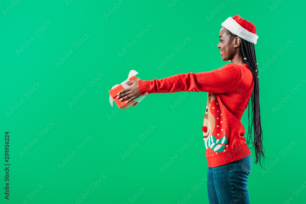 side view of African American woman in Christmas sweater and Santa hat raising hands with gift box isolated on green
