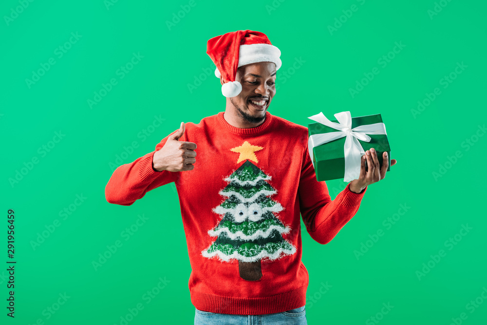African American man in Christmas sweater and Santa hat showing thumb up and looking at gift box in his hands isolated on green African American man in Christmas sweater and Santa hat showing thumb up and looking at gift box in his hands isolated on green