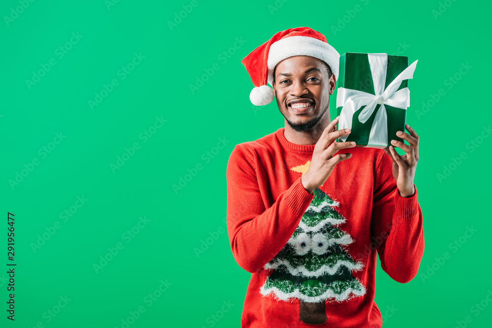 African American man in Christmas sweater and Santa hat holding present and  looking at camera isolated on green Stock Photo | Adobe Stock
