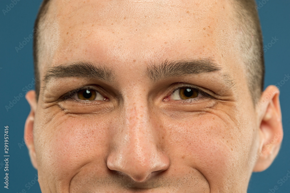 Fototapeta premium Caucasian young man's close up shot on blue studio background. Beautiful model with well-kept skin. Concept of human emotions, facial expression, male beauty and healthcare. Eyes smiling and eyebrow.