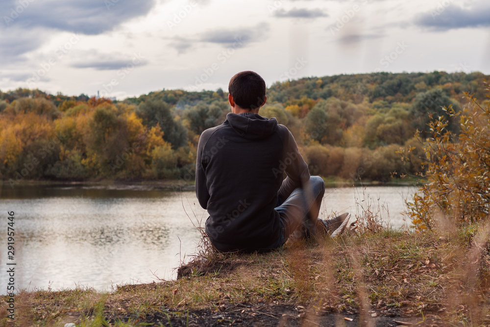 A young man sits on the Bank of the river autumn cloudy evening. Back view