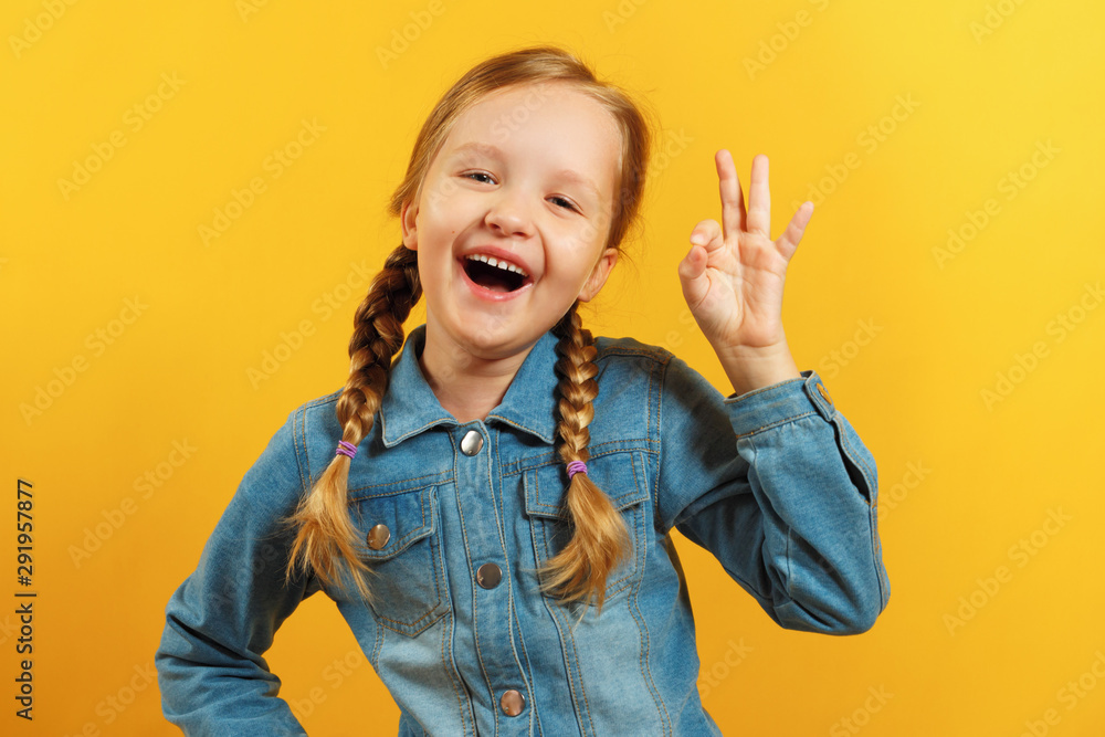 Little girl in a denim shirt on a yellow background. The child shows an OK sign. The concept of success, approval Little girl in a denim shirt on a yellow background. The child shows an OK sign. The concept of success, approval