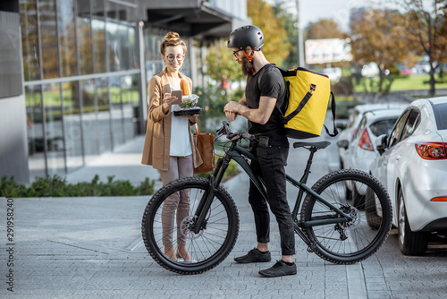 Courier delivering fresh lunches to a young business woman on a bicycle with thermal backpack. Takeaway restaurant food delivery concept