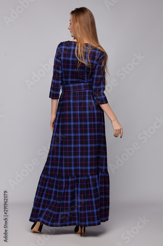 Back view of young blonde European lady with long straight hair wearing a black maxi dress with a print Scottish tartane, with three-quarter sleeves, a wide frill along the hem and a belt.   photo