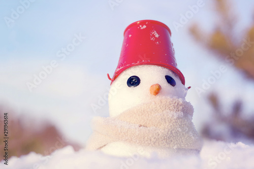 Little snowman made of snow in scarf and red bucket on his head on snow on blue sky background, soft focus. Christmas background © isavira