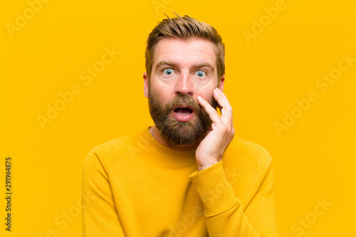 young blonde man open-mouthed in shock and disbelief, with hand on cheek and arm crossed, feeling stupefied and amazed against orange wall photo