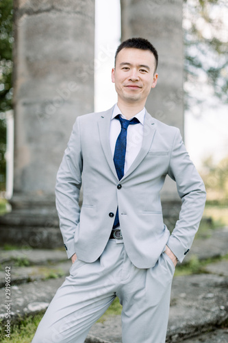 Elegant smiling handsome Asian groom in gray suit posing on the background of ancient stone columns in old castle
