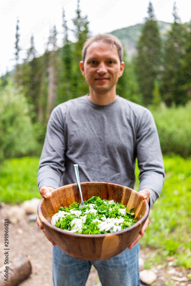 Young man holding kale salad large wooden bowl with background of green Utah mountains in Albion Basin campground Young man holding kale salad large wooden bowl with background of green Utah mountains in Albion Basin campground