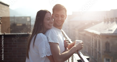 Handsome young male in t-shirt drinking coffee on balcony, loving wife hugging him, sweet relations, love