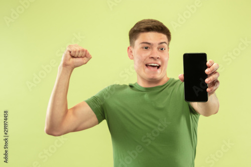 Caucasian young man's half-length portrait on green studio background. Beautiful male model in shirt. Concept of human emotions, facial expression, ad. Showing phone's screen, celebrating, happy.