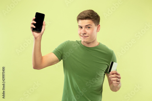 Caucasian young man's half-length portrait on green studio background. Beautiful male model in shirt. Concept of human emotions, facial expression, sales, ad. Holding phone and card, online payments.
