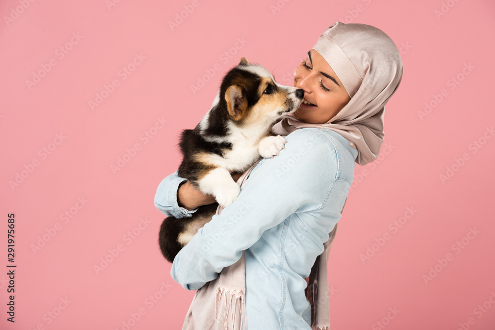 arabic girl in hijab holding cute corgi puppy, isolated on pink