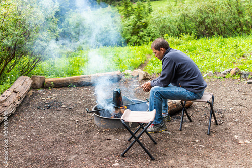 Campsite with man sitting on chair warming hands by firepit smoke and teapot on grill in Albion Basin, Utah summer in Wasatch mountains in evening