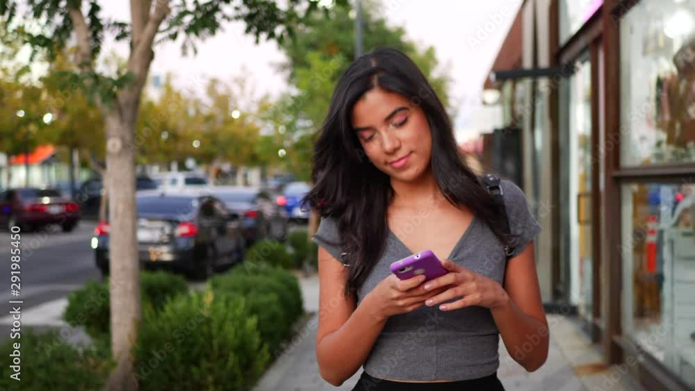 A young hispanic woman walking and smiling on an urban city street sidewalk texting on her smartphone. A young hispanic woman walking and smiling on an urban city street sidewalk texting on her smartphone.
