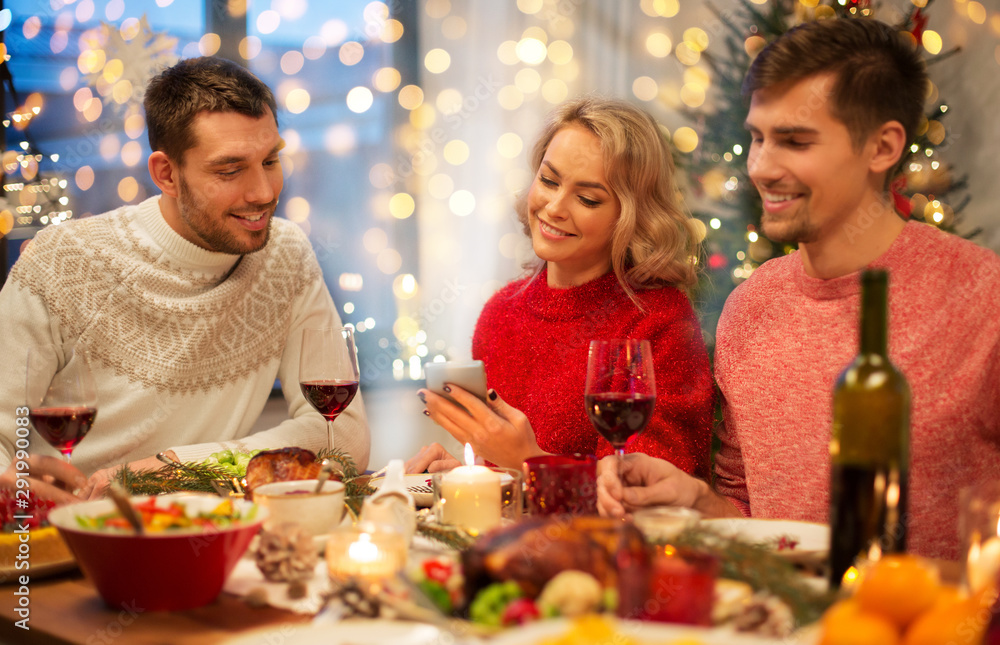 holidays and celebration concept - happy friends with smartphone having christmas dinner at home holidays and celebration concept - happy friends with smartphone having christmas dinner at home