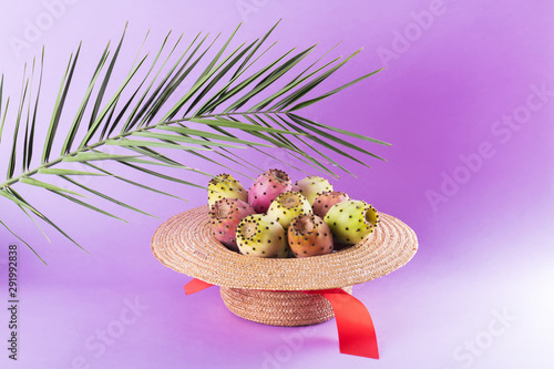 Opuntia fruit in a straw hat with a palm leaf on a trendy purple background. Prickly pear or Fichi Dindia Salento, Puglia. Minimalism, pop art photo