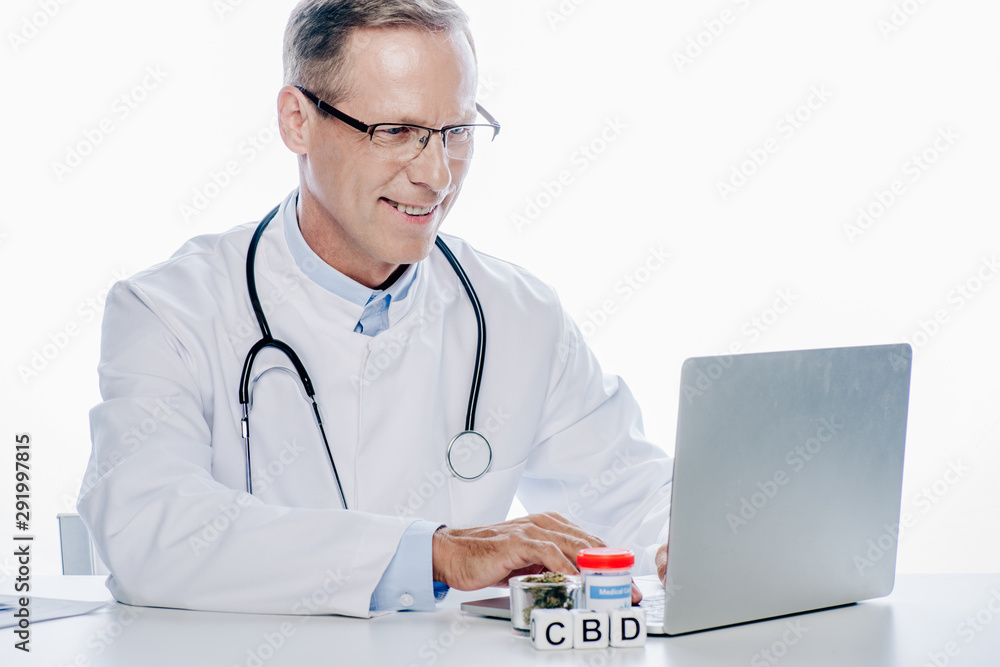 handsome and smiling doctor in white coat using laptop isolated on white handsome and smiling doctor in white coat using laptop isolated on white