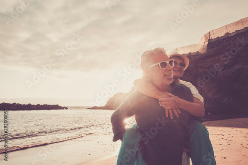 beautiful couple of seniors at the beach having fun together and playing - mature man carry his wife woman with love and forever life concept - sea or ocean in the background -   retired lifestyle