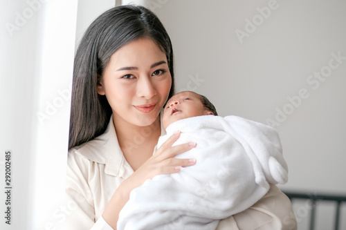 Beautiful Asian mother hold her newborn baby touch her face, smile and look forward in front of glass window with day light.