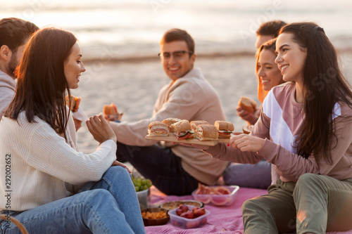 friendship  leisure and fast food concept - group of happy friends eating sandwiches or burgers at picnic on beach in summer