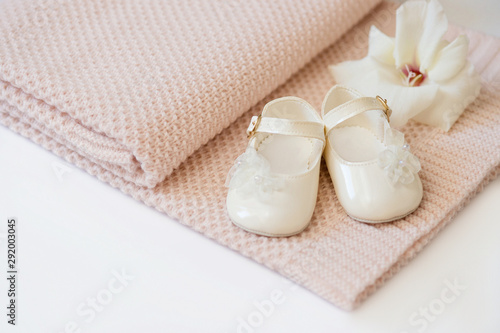 Pink knitted plaid lies on a white surface. White baby boots and a white gladiolus flower lie on it.
