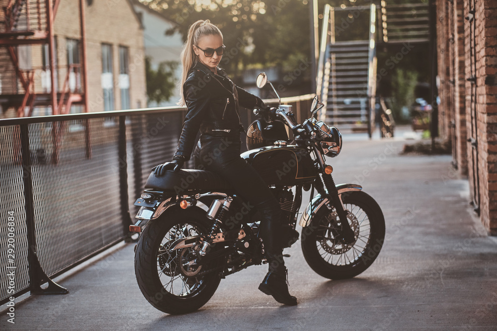 Stylish female biker is sitting at her black bike while posing for a photoshoot on the street.