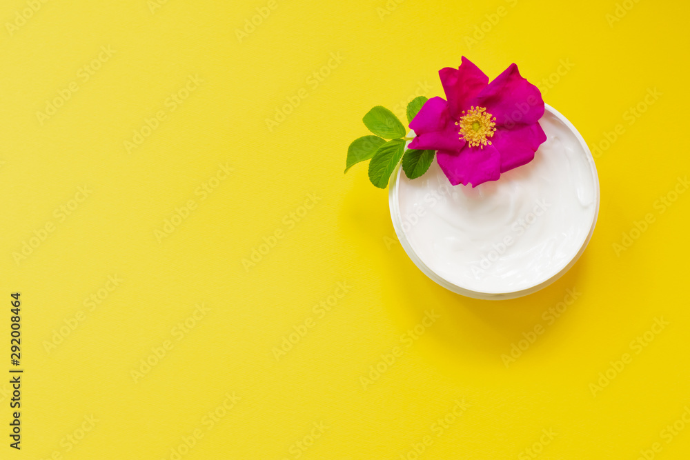 White soft cream in a jar on bright yellow background with dog rose, top view, anti-ageing moisturizing cream with dog rose oil essential and vitamin E