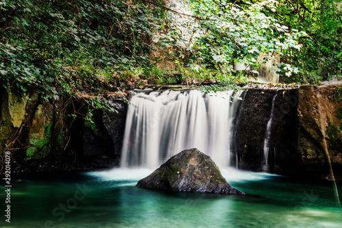 Small waterfall in the middle of nature  which forms a pond with a rock in the middle  green woods  leaves  roots and climbing plants.