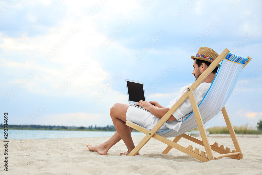 Young man with laptop sitting in deck chair on sea beach. Space for text
