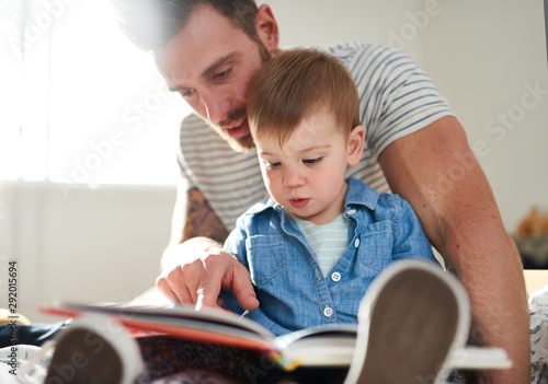 Adorable, young female toddler learning to read book with handsome millennial father on bed