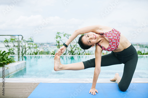 Portrait of Asian young woman smiling at camera while warming up on exercise mat before training near the poolside outdoors