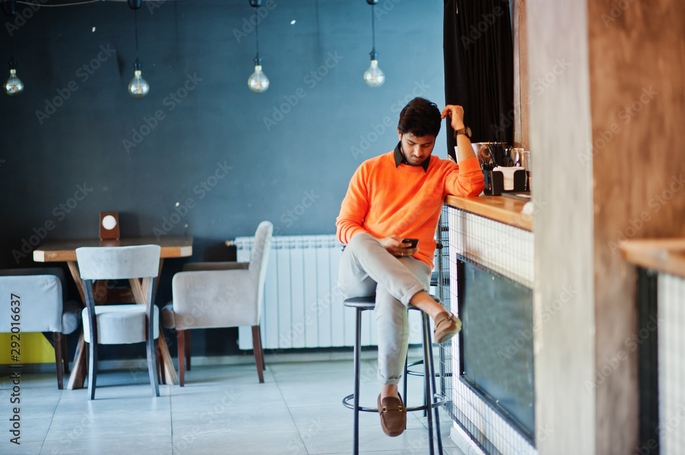 Confident young indian man in orange sweater sitting bar counter at cafe with mobile phone at hand.