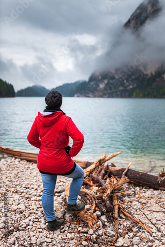 Women with red rain jacket standing all alone on a rocky mountain lake bank on a cloudy misty rainy autumn day and enjoying the peacful nature view after a hike.